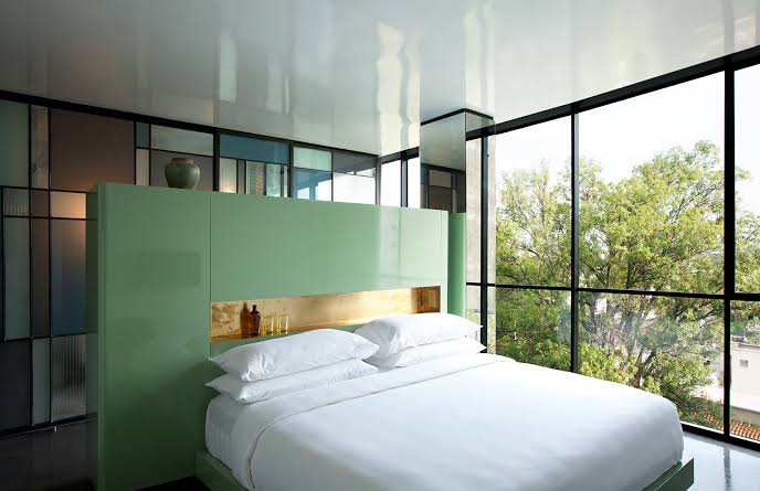 A bedroom with a green headboard, white bedding and a window onto trees.