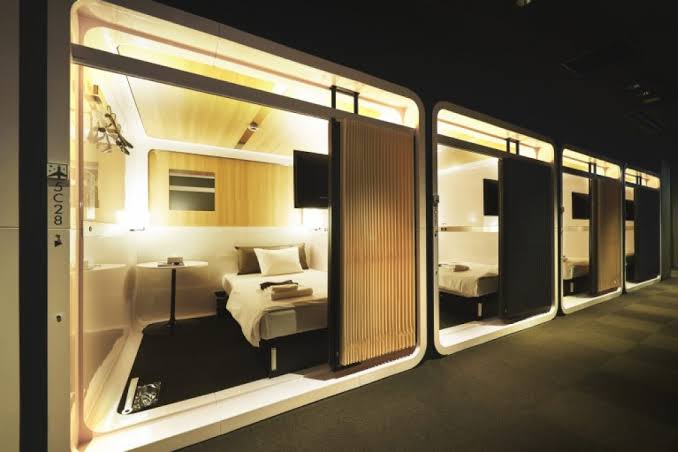 A row of cabins along a corridor, one open to show a bed and a small table.