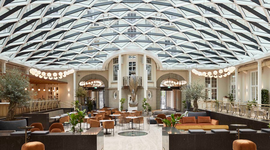 A glass-roofed atrium with cafe tables, orange seating and hanging globe lights.
